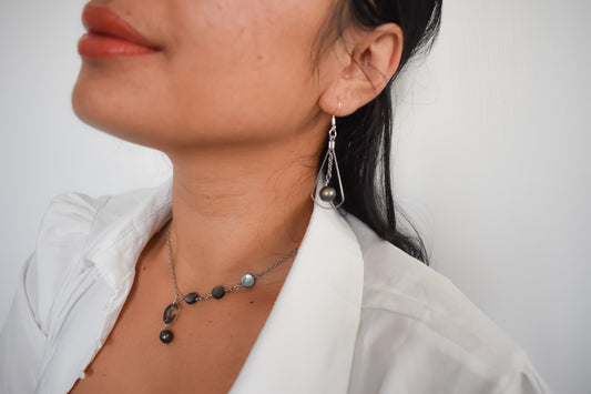 Une femme portant un collier en fin argenté en acier inoxydable, orné de trois petites nacres rondes et d’un pendentif lune en nacre accompagné d’une petite perle de Tahiti, assorti à une boucle d’oreille élégante, sur fond blanc