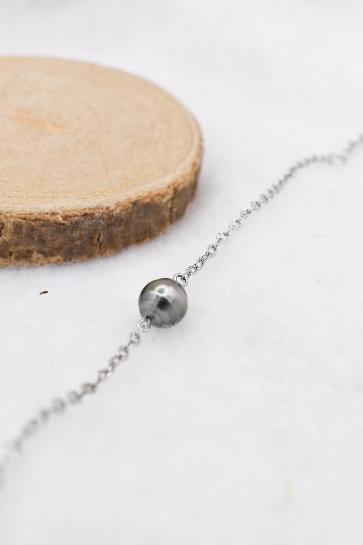 Bracelet minimaliste en chaine fine acier inoxydable et sa petite perle de Tahiti, posé sur un fond blanc et près d’une rondelle de bois naturelle.