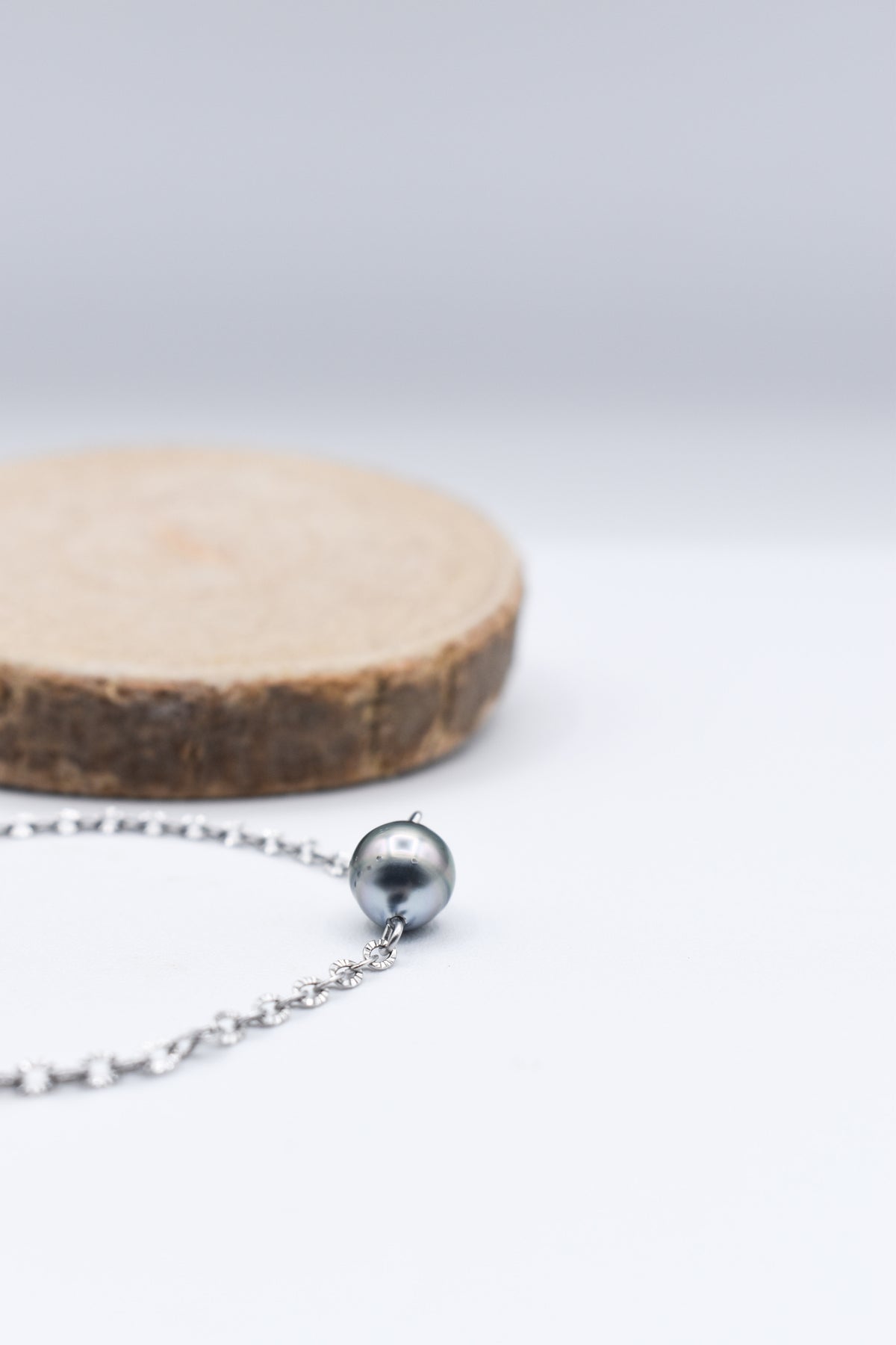 Bracelet en chaîne fine avec sa perle de Tahiti qui brille de ces belles couleurs irisée posé sur un fond blanc près d’un rondelle de bois naturel